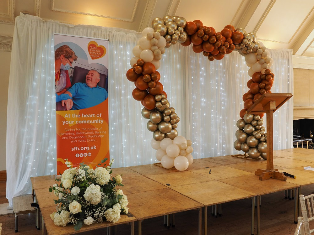 Stage set with heart shape balloons and pop up poster for Saint Francis Hospice, beautiful big bouquet of white flowers on the floor_Big Heart Lunch, event for Saint Francis Hospice